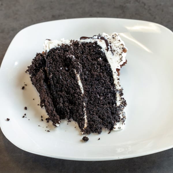 A slice of chocolate cake with white frosting and chocolate drizzle on a white plate.