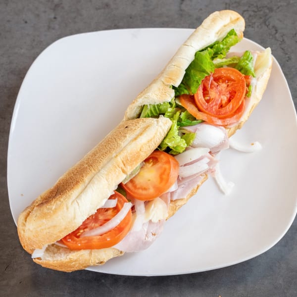 A long submarine sandwich, overflowing with ham, lettuce, tomatoes, and onions, on a white plate.